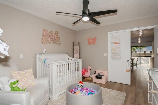 bedroom with ceiling fan, a nursery area, dark wood-type flooring, and a textured ceiling