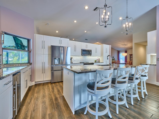 kitchen featuring appliances with stainless steel finishes, white cabinets, dark stone counters, hanging light fixtures, and a center island with sink