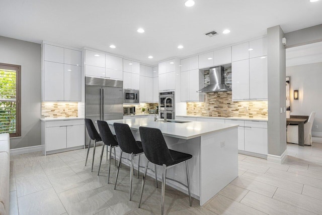 kitchen with wall chimney range hood, white cabinetry, built in appliances, and an island with sink