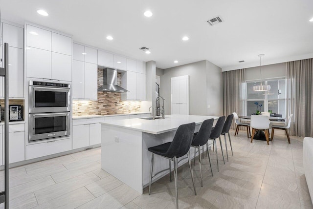 kitchen with white cabinetry, a kitchen breakfast bar, wall chimney exhaust hood, a center island with sink, and stainless steel double oven