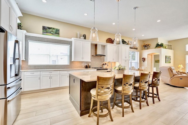 kitchen with appliances with stainless steel finishes, a kitchen island with sink, hanging light fixtures, white cabinets, and wall chimney exhaust hood