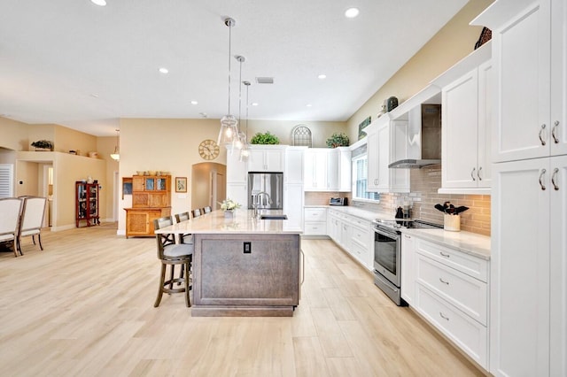 kitchen with an island with sink, white cabinets, hanging light fixtures, stainless steel appliances, and wall chimney range hood