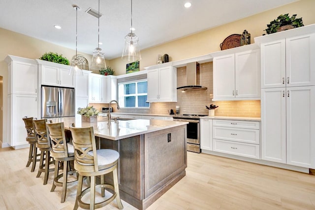 kitchen featuring appliances with stainless steel finishes, pendant lighting, white cabinets, wall chimney range hood, and a center island with sink