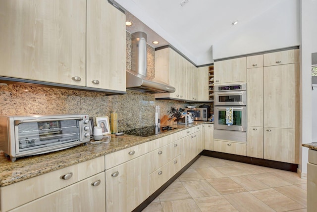 kitchen featuring lofted ceiling, wall chimney range hood, light stone countertops, light brown cabinetry, and stainless steel double oven