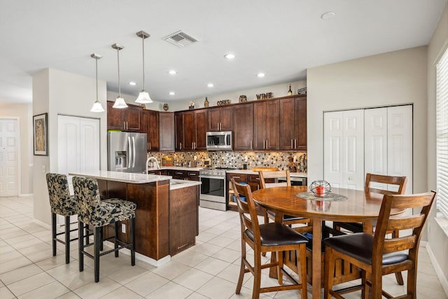 kitchen with decorative backsplash, hanging light fixtures, a kitchen island with sink, dark brown cabinetry, and stainless steel appliances