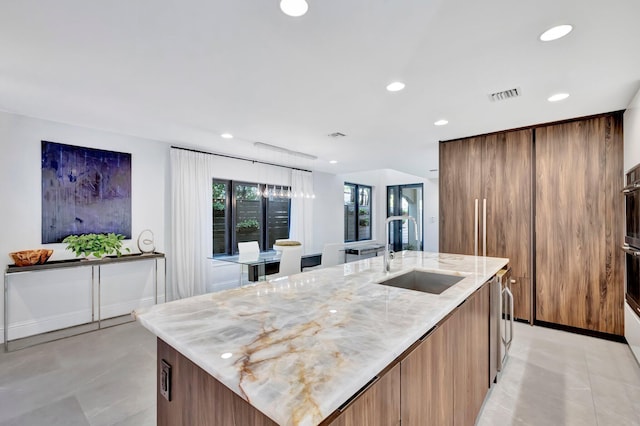 kitchen with light stone counters, stainless steel oven, sink, and a center island with sink