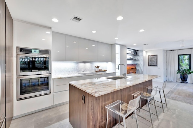 kitchen featuring sink, a breakfast bar area, light stone countertops, a kitchen island with sink, and white cabinets