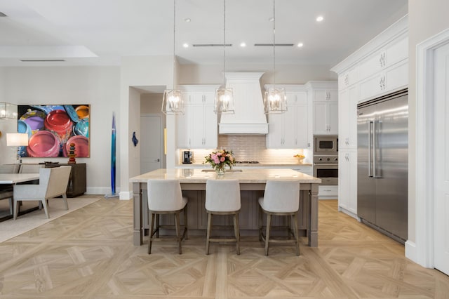 kitchen featuring pendant lighting, built in appliances, custom range hood, white cabinets, and a center island with sink