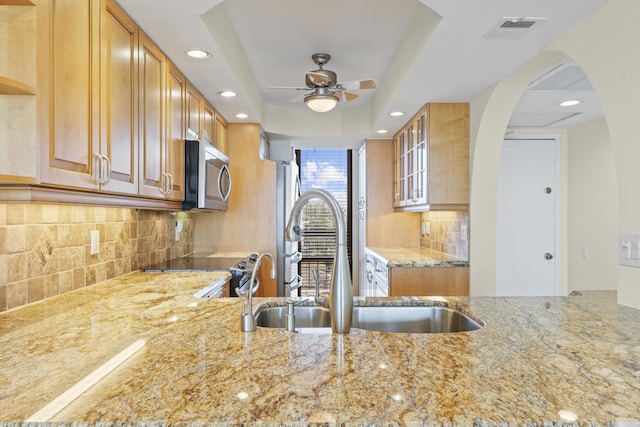 kitchen featuring sink, ceiling fan, a tray ceiling, light stone countertops, and decorative backsplash