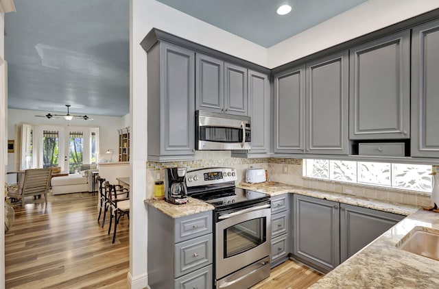 kitchen with gray cabinetry, backsplash, stainless steel appliances, light stone counters, and light wood-type flooring