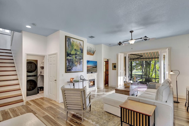 living room featuring ceiling fan, stacked washer and clothes dryer, light hardwood / wood-style flooring, and a textured ceiling