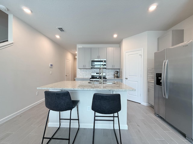 kitchen featuring gray cabinets, backsplash, stainless steel appliances, light stone counters, and an island with sink