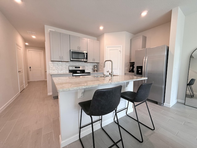 kitchen featuring sink, appliances with stainless steel finishes, light stone countertops, an island with sink, and decorative backsplash