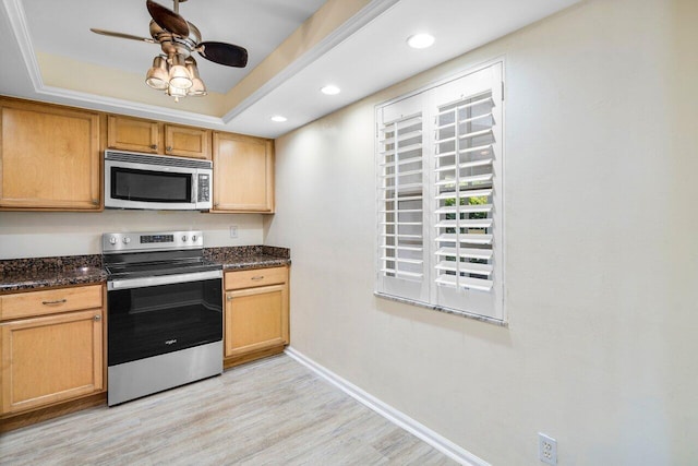 kitchen with ceiling fan, dark stone countertops, stainless steel appliances, a tray ceiling, and light wood-type flooring