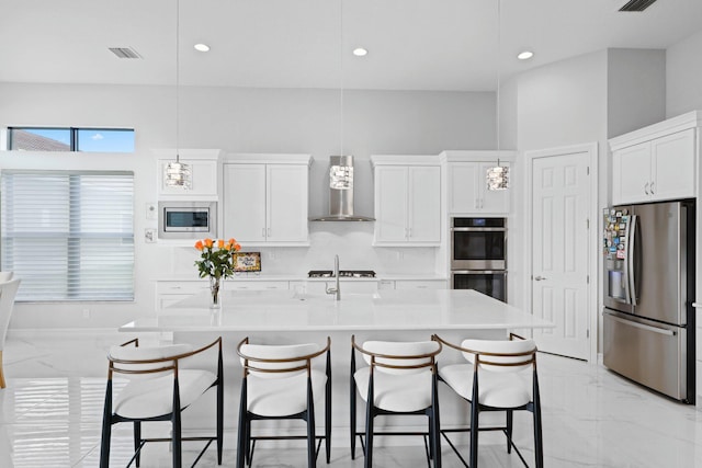 kitchen with decorative light fixtures, white cabinetry, wall chimney range hood, stainless steel appliances, and a center island with sink