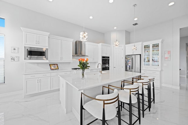 kitchen with white cabinetry, appliances with stainless steel finishes, an island with sink, pendant lighting, and wall chimney range hood