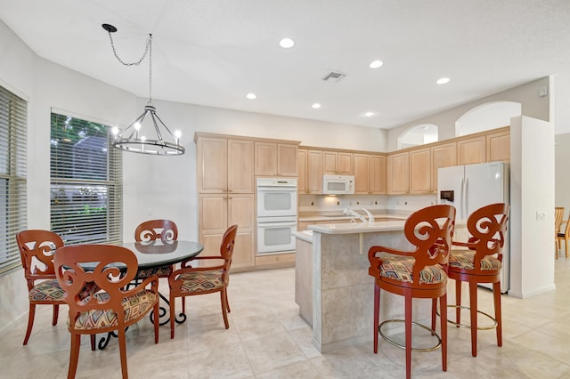 kitchen with pendant lighting, an island with sink, a chandelier, light brown cabinets, and white appliances