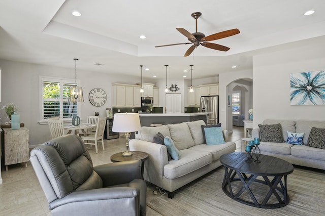 living room with light tile patterned floors, ceiling fan with notable chandelier, and a raised ceiling