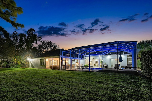 back house at dusk featuring a patio, glass enclosure, and a lawn