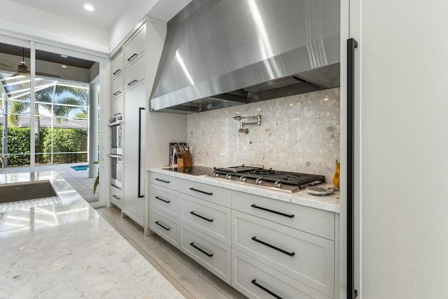 kitchen with appliances with stainless steel finishes, white cabinets, backsplash, light stone counters, and wall chimney range hood