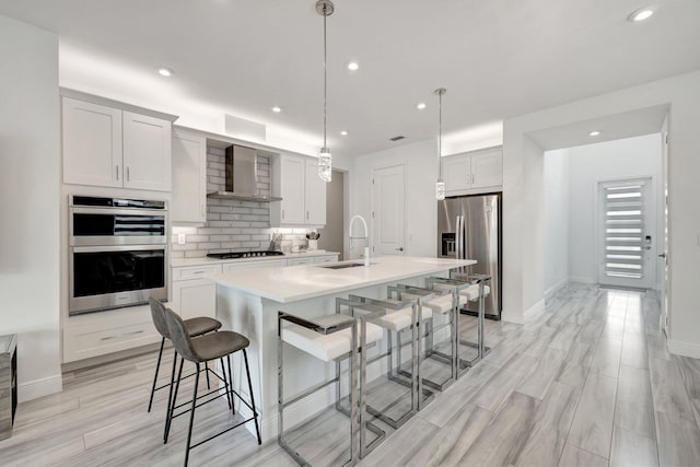 kitchen featuring sink, hanging light fixtures, an island with sink, stainless steel appliances, and wall chimney range hood