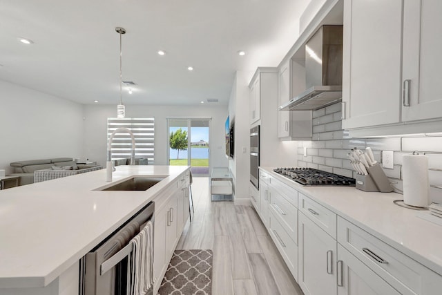 kitchen featuring sink, wall chimney range hood, appliances with stainless steel finishes, white cabinetry, and hanging light fixtures