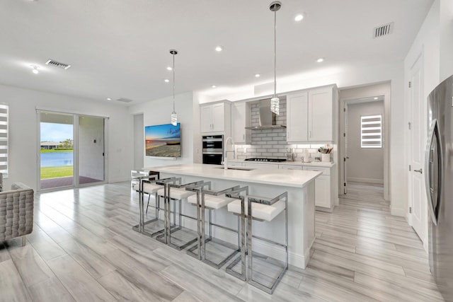 kitchen with pendant lighting, a kitchen island with sink, stainless steel appliances, white cabinets, and wall chimney exhaust hood