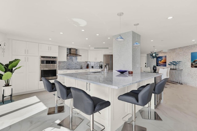 kitchen with appliances with stainless steel finishes, white cabinetry, hanging light fixtures, light stone counters, and wall chimney exhaust hood