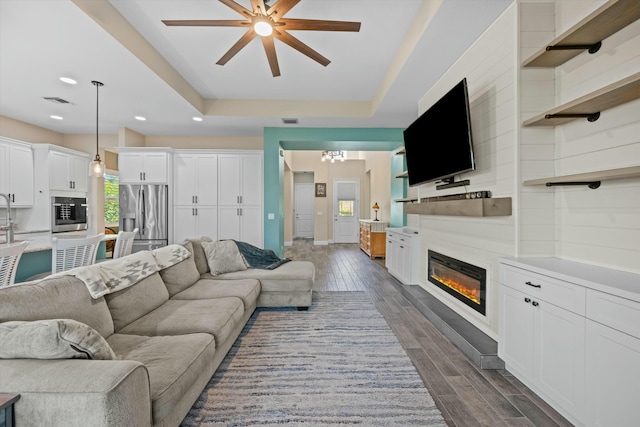 living room featuring a tray ceiling, dark wood-type flooring, a large fireplace, and ceiling fan