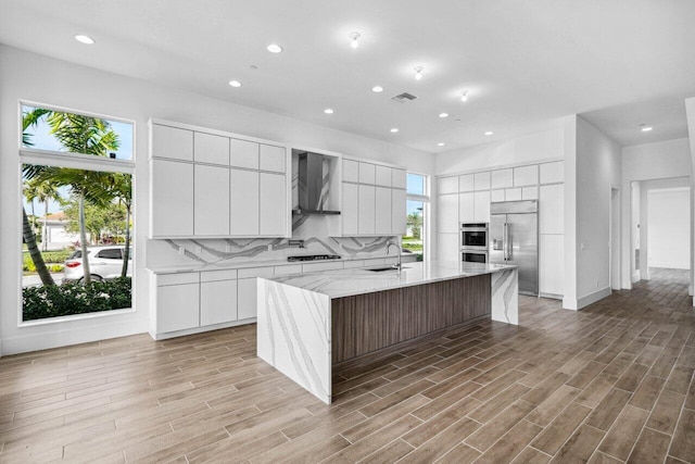 kitchen with a spacious island, wall chimney range hood, stainless steel appliances, decorative backsplash, and white cabinets