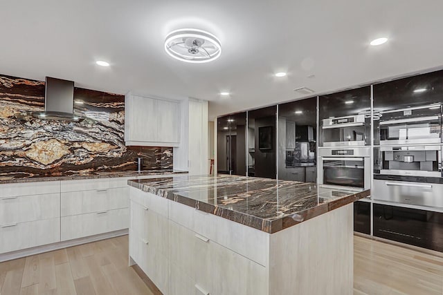 kitchen with a spacious island, wall chimney exhaust hood, light hardwood / wood-style flooring, stainless steel double oven, and dark stone counters