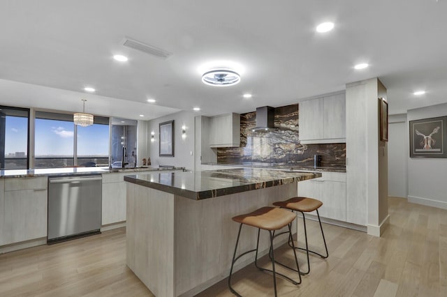 kitchen featuring pendant lighting, dark stone countertops, stainless steel dishwasher, wall chimney exhaust hood, and light wood-type flooring