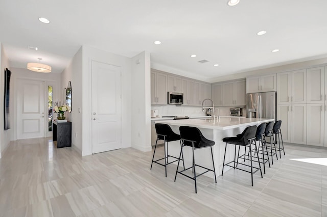 kitchen with stainless steel appliances, a kitchen island with sink, gray cabinetry, and a kitchen bar