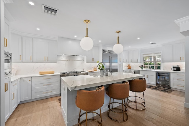 kitchen with white cabinetry, built in appliances, hanging light fixtures, an island with sink, and beverage cooler