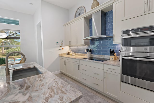 kitchen featuring sink, double oven, light stone counters, black electric cooktop, and wall chimney exhaust hood