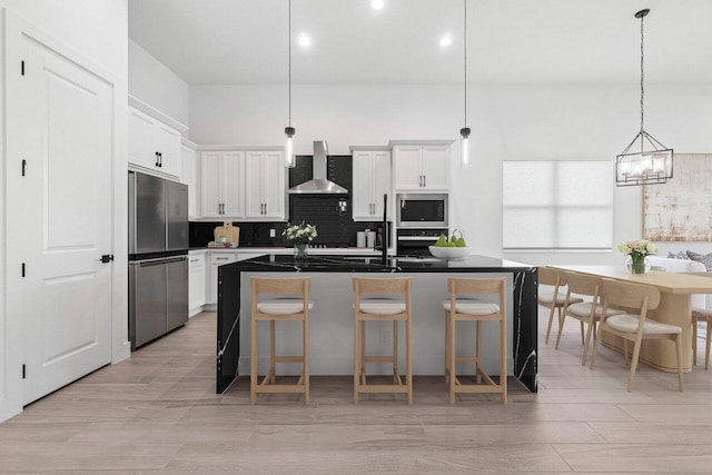 kitchen featuring white cabinetry, wall chimney range hood, a kitchen island with sink, and appliances with stainless steel finishes