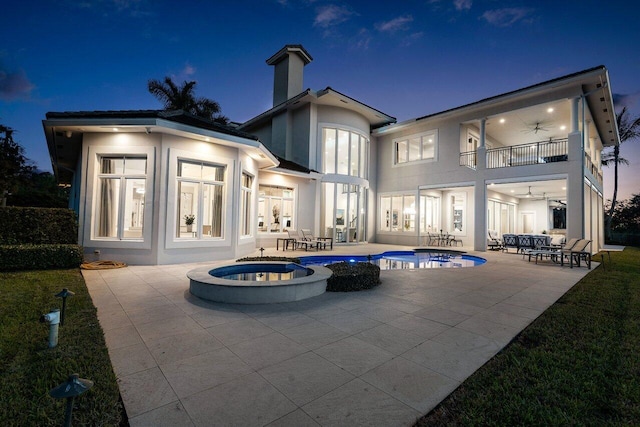 back house at twilight with a swimming pool with hot tub, a patio area, a balcony, and ceiling fan