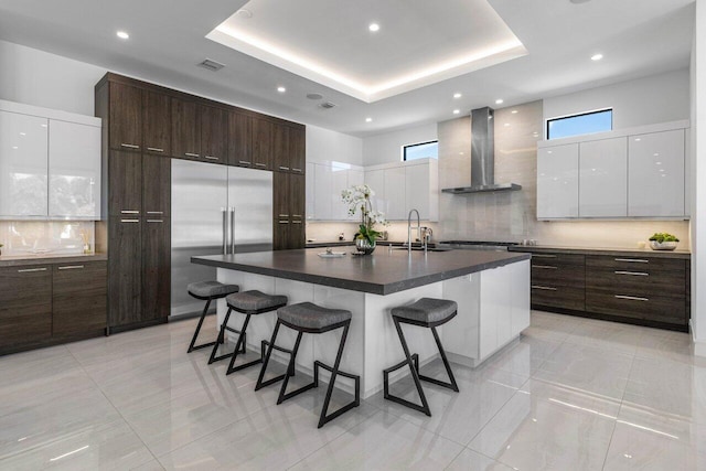 kitchen featuring modern cabinets, stainless steel built in refrigerator, a sink, a tray ceiling, and wall chimney exhaust hood