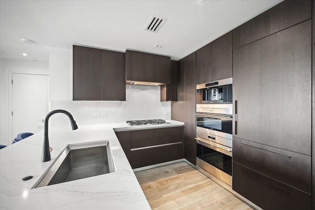 kitchen with appliances with stainless steel finishes, sink, light stone counters, dark brown cabinets, and light wood-type flooring