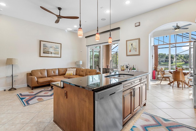 kitchen with light tile patterned flooring, dark stone counters, hanging light fixtures, a kitchen island with sink, and stainless steel dishwasher