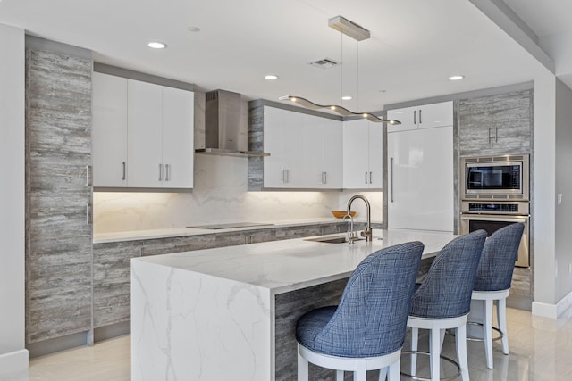 kitchen with pendant lighting, an island with sink, sink, stainless steel appliances, and wall chimney exhaust hood