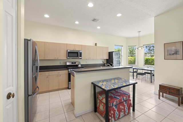 kitchen featuring light brown cabinetry, decorative light fixtures, a center island with sink, light tile patterned floors, and appliances with stainless steel finishes