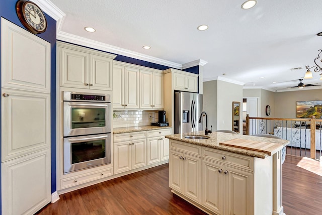 kitchen with sink, a kitchen island with sink, stainless steel appliances, cream cabinets, and light stone countertops