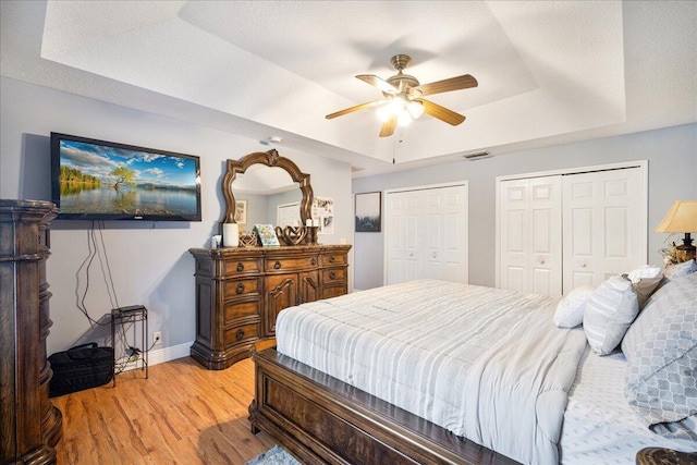 bedroom featuring ceiling fan, a tray ceiling, light hardwood / wood-style floors, a textured ceiling, and two closets