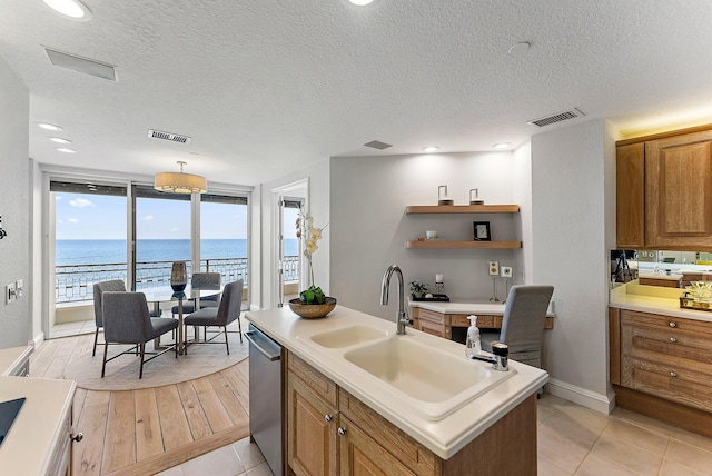 kitchen with sink, a water view, a textured ceiling, stainless steel dishwasher, and an island with sink