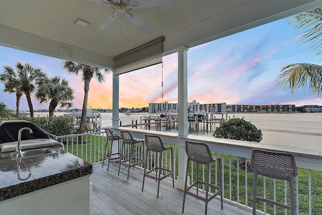 deck at dusk featuring a water view, ceiling fan, a grill, and a bar