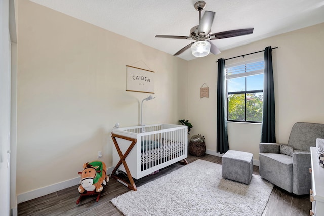 bedroom with a nursery area, dark wood-type flooring, and ceiling fan