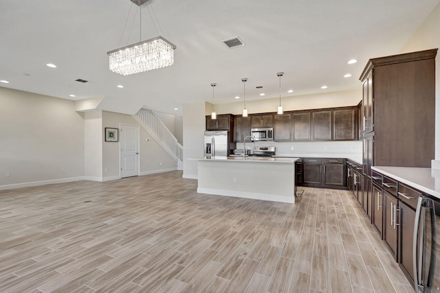 kitchen with light hardwood / wood-style flooring, appliances with stainless steel finishes, a kitchen island with sink, hanging light fixtures, and dark brown cabinetry