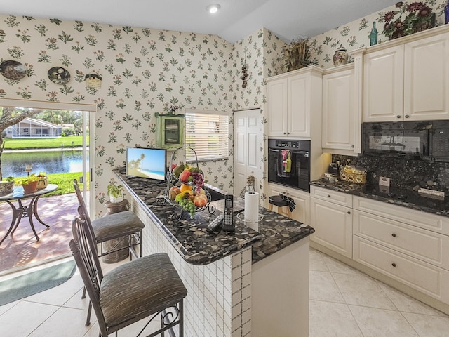 kitchen with light tile patterned flooring, lofted ceiling, a breakfast bar, black oven, and dark stone counters