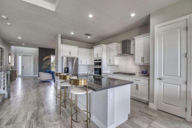 kitchen featuring a breakfast bar area, wall chimney exhaust hood, stainless steel appliances, white cabinets, and a kitchen island with sink